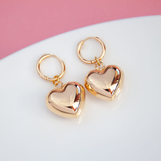 Gold heart-shaped earrings on a white surface with a pink background